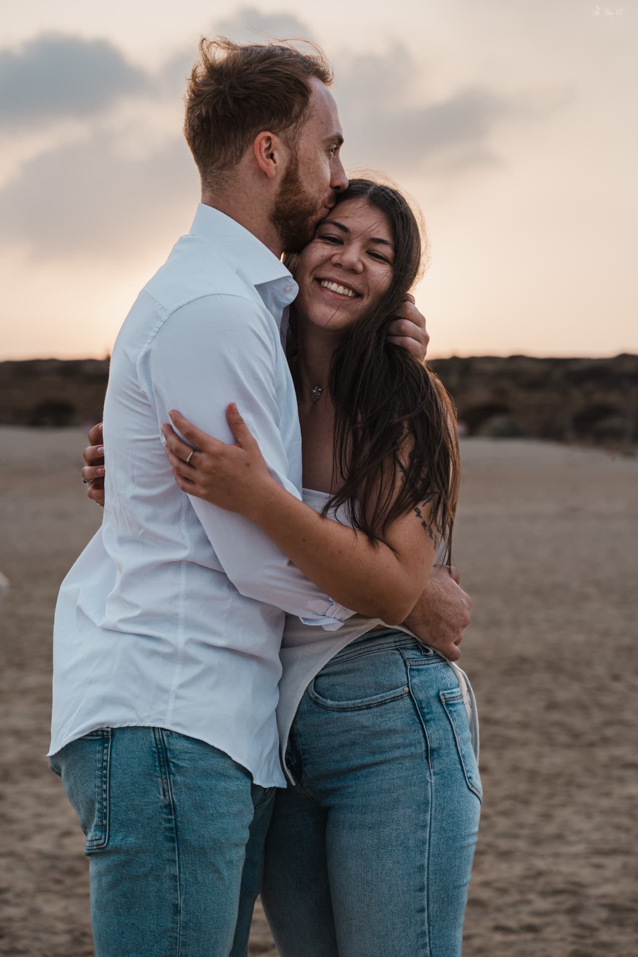זוג מחובק על החוף בשקיעה, הוא מנשק את ראשה והיא מחייכת למצלמה